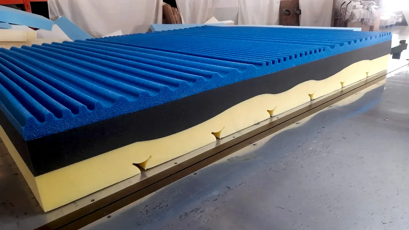 Materasso su misura a più strati: poliuretano giallo con taglio a onda, strato grigio e memory foam blu bugnato sul banco di taglio